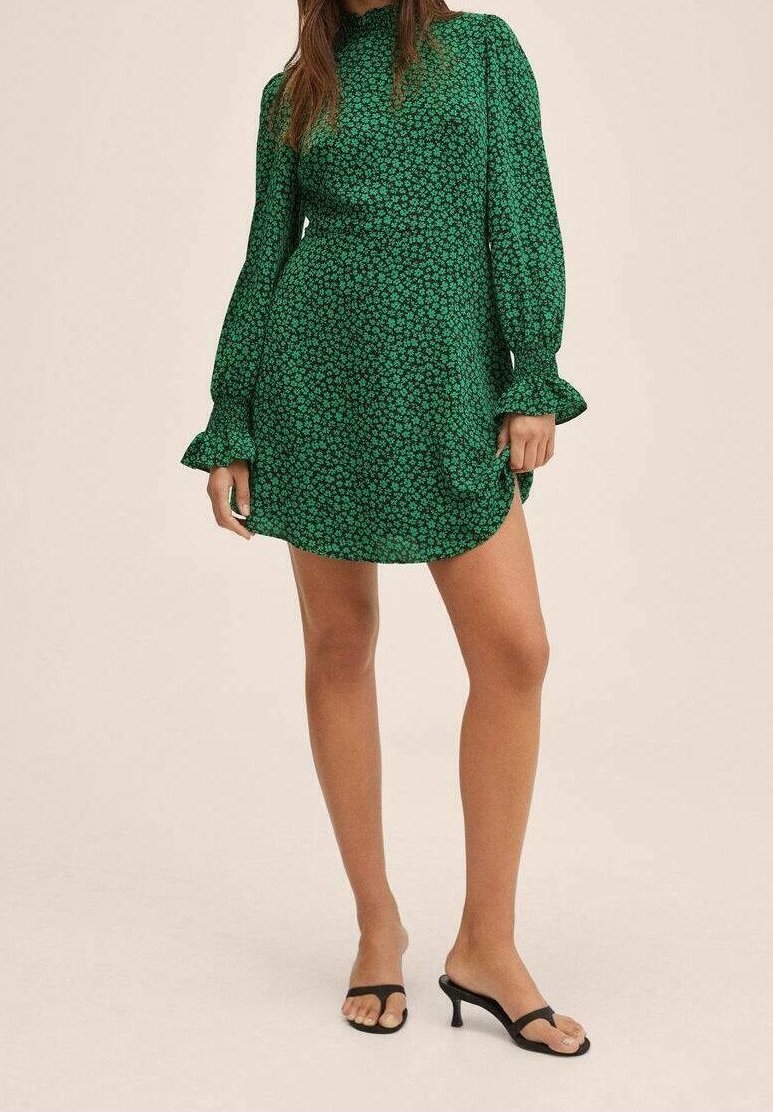 Robe florale verte avec de longues manches bouffantes, un col haut et des poignets volants. Présente une forme évasée et un tissu léger. Portée avec des sandales noires.