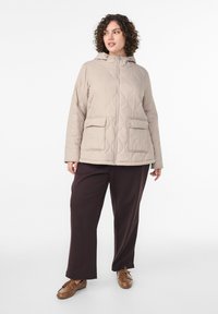 Beige quiltet jakke med hætte, der har to forlommer og lynlås. Kompletteret med mørkebrune bukser med vide ben og brune sko.