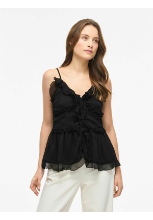 Žena s dugom smeđom kosom, odjevena u crnu bluzu bez rukava s naborima i bijele hlače, stoji ispred jednobojne bijele pozadine.