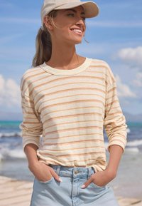 Stribet langærmet top i lys creme med orange og gule striber, parret med lyseblå denim shorts. Uformel strandbaggrund.