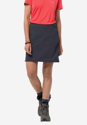 Femme portant une chemise corail à manches courtes, une jupe bleu marine jusqu'aux genoux, des bottes de randonnée et un bracelet, debout avec une jambe légèrement croisée.