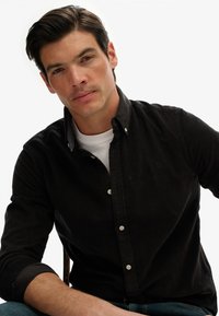 Camisa de corduroy negra con botones y un leve acento en el collar, usada sobre una camiseta blanca. El modelo tiene el pelo corto y oscuro y está sentado de manera casual.