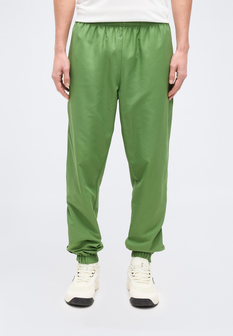 Άτομο που φοράει πράσινα παντελόνια τζόγκερ με ελαστική μέση και εκρού αθλητικά παπούτσια, στέκεται μπροστά σε ένα απλό λευκό φόντο.