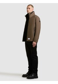 Brązowa pikowana kurtka z wysokim kołnierzem, boczne kieszenie; noszona na czarnej bluzce i czarnych spodniach, w zestawie z czarnymi sznurowanymi butami.