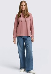 Rózsaszín kapucnis pulóver húzózsinórokkal, laza szabású, világoskék bőszárú farmernadrággal és fehér tornacipővel párosítva, semleges háttér előtt állva.