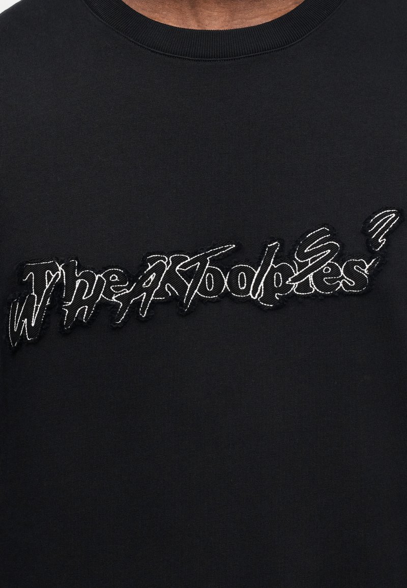 Sweatshirt noir avec texte brodé "TheAdooples?" en blanc dans une police irrégulière et dentelée. Encolure côtelée et tissu texturé.