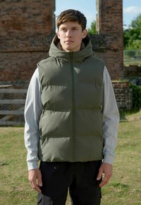 Grüne Daunenweste mit Kapuze, mit glatter Textur und Vorderreißverschluss. Getragen über einem hellgrauen Langarmshirt und dunklen Hosen.