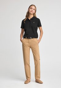 Polo noir à manches courtes, avec un plastron à un seul bouton et un petit logo, associé à un chino beige et des mocassins marron clair. Texture lisse.