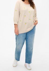 Pullover lavorato a maglia beige chiaro con ricamo di ananas giallo, abbinato a jeans in denim blu larghi e sneakers bianche con dettagli rosa.