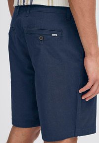 Shorts bleu marine en tissu léger, dotés d'une poche arrière avec fermeture à bouton et d'une étiquette de marque discrète. Texture lisse.