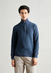 Dunkelblauer gerippter Strickpullover mit hohem Kragen und einem halbseitigen Reißverschluss. Verfügt über einen strukturierten Stoff und eng anliegende Bündchen. Getragen über hellen Hosen.