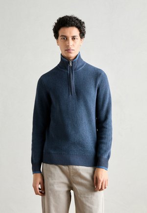 Dunkelblauer gerippter Strickpullover mit hohem Kragen und einem halbseitigen Reißverschluss. Verfügt über einen strukturierten Stoff und eng anliegende Bündchen. Getragen über hellen Hosen.