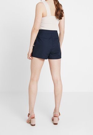 Kvinde iført ærmeløs beige top, marineblå højtaljede shorts med knapdetaljer og beige sandaler med blokhæl, stående mod hvid baggrund.