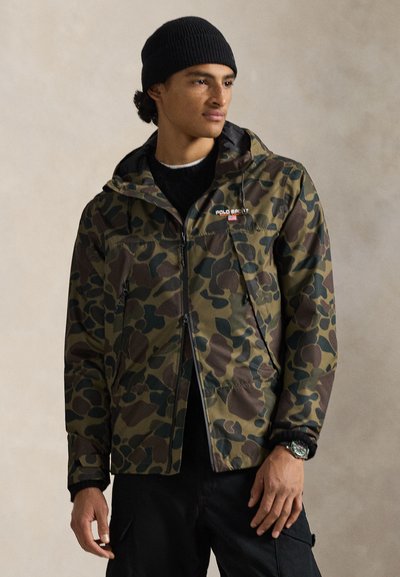 Chaqueta con patrón de camuflaje en verde y marrón, con capucha, bolsillos con cremallera y puños ajustables. Usada sobre una camisa negra.