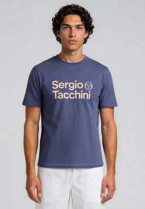 Ung mand med mørkt krøllet hår iført en blå "Sergio Tacchini" t-shirt og hvide shorts mod en ensfarvet grå baggrund.