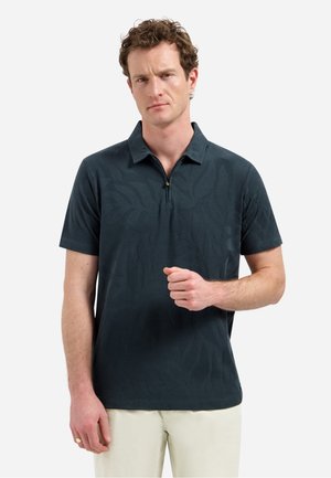 Man met krullend haar, gekleed in een donkerblauw poloshirt met korte mouwen en een subtiel bladmotief, en beige broek, staand tegen een witte achtergrond.