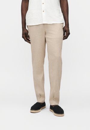 Mężczyzna w beżowych prostych spodniach, białej koszuli na guziki i czarnych espadrylach na jednolitym tle.