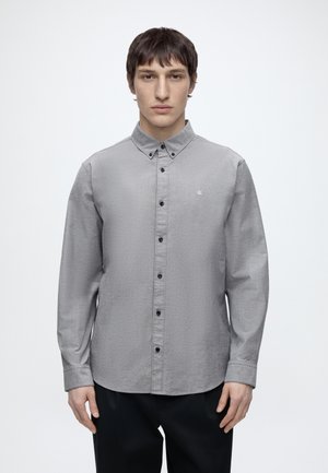 Jeune homme de face, portant une chemise grise à boutons noirs avec un petit logo sur la poitrine, debout devant un fond blanc uni.
