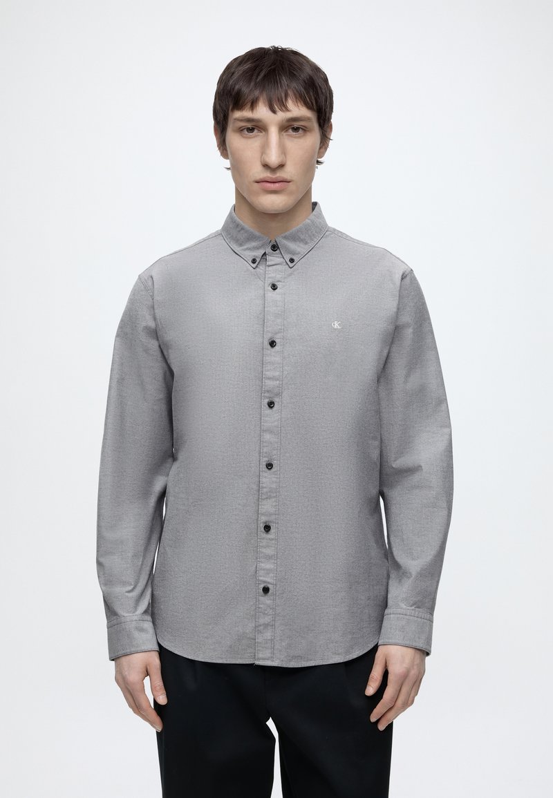 Jeune homme de face, portant une chemise grise à boutons noirs avec un petit logo sur la poitrine, debout devant un fond blanc uni.