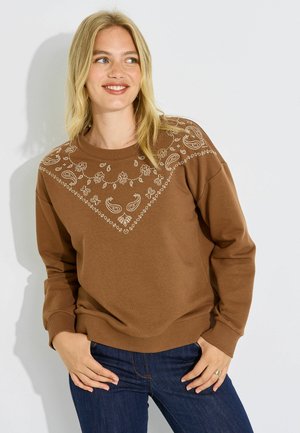 Mujer rubia con una sudadera marrón con patrones blancos de paisley en el pecho, posando con las manos en los bolsillos de unos vaqueros azules.