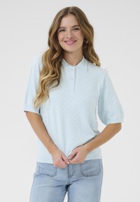 Helles blaues Polo-Sweatshirt mit strukturiertem Rautenmuster, kurzen Ärmeln und einer Knopfleiste mit drei Knöpfen. Kombiniert mit hellen Denim-Jeans.