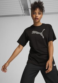 Černé bavlněné tričko s texturovaným bílým logem Puma ve designu skákajícího kočky. Krátké rukávy, kulatý výstřih, uvolněný střih, nošené s cargo kalhotami.