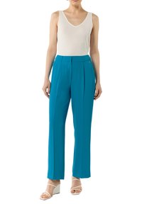 Jasnozielone dopasowane spodnie z zakładkami, wysoki stan i prosty krój, w połączeniu z białym bezrękawnikem i białymi sandałami na słupku.