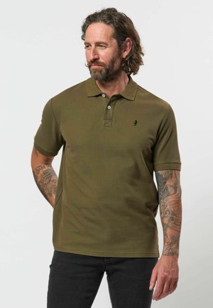 Mann mit Bart und Tattoos trägt ein olivgrünes Poloshirt und schwarze Hose, steht mit den Händen hinter dem Rücken und schaut zur Seite.