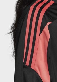 Svart jacka med rosa och svarta randiga detaljer på axeln. Glatt tyg med kontrasterande tygpaneler. Närbild.