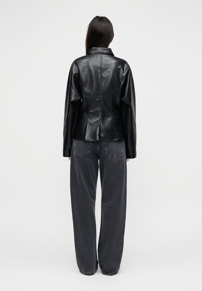 Mustast nahast jakk, millel on struktureeritud siluett, volditud krae ja pikad varrukad, kombineeritud lahtiste tumedate hallide teksapükstega.