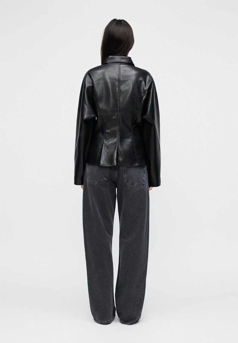 Veste en cuir noire à silhouette structurée, avec un col plié et des manches longues, associée à un pantalon en denim gris foncé à coupe ample.