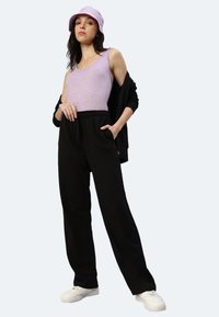 Hellblaues ärmelloses Oberteil, schwarze, locker sitzende Hose mit elastischem Bund, schwarze Strickjacke und lilafarbener Bucket Hat. Weiße Sneakers.