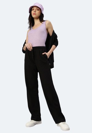 Hellblaues ärmelloses Oberteil, schwarze, locker sitzende Hose mit elastischem Bund, schwarze Strickjacke und lilafarbener Bucket Hat. Weiße Sneakers.