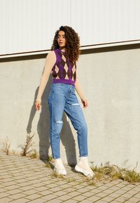 Colete de argyle em roxo e castanho sobre uma camisa de manga longa bege, combinado com jeans azuis desgastados e ténis de cano alto bege claro.