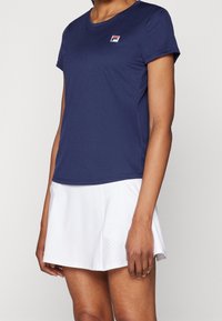 Námorně modré sportovní tričko s krátkými rukávy a kulatým výstřihem, se malým červeným a bílým logem. Nosené s bílou perforovanou sukní.