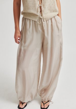Persona che indossa pantaloni beige larghi con coulisse abbinati a un gilet senza maniche a maglia e infradito nere, in piedi contro uno sfondo semplice.