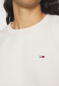 Beige Sweatshirt mit Rundhalsausschnitt, strukturiertem Stoff und kleinem gesticktem Logo auf der Brust mit roten, weißen und blauen Details.