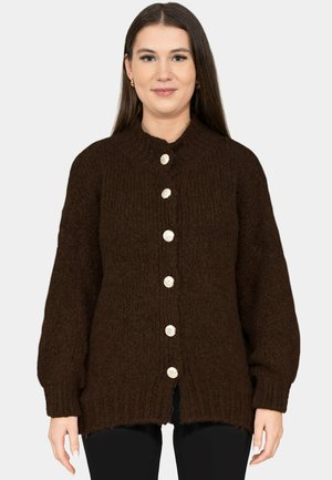 Femme portant un cardigan en maille épaisse marron foncé avec de gros boutons argentés, un pantalon noir et des boucles d'oreilles créoles dorées, sur un fond uni.