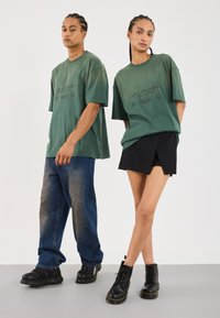 Grønne oversized t-shirts med fronttrykt, parret med mørke shorts. Modellerne bærer sorte støvler og sneakers. Ensfarvet baggrund, minimale forstyrrelser.