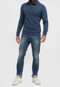 Niebieski dziany sweter z wysokim kołnierzem i ozdobnymi guzikami, w zestawieniu z jasnobłękitnymi dżinsami i białymi trampkami.