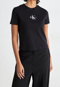 Camiseta de algodón negra corta con cuello redondo y un logo de Calvin Klein impreso en blanco en el lado izquierdo del pecho. Textura suave.