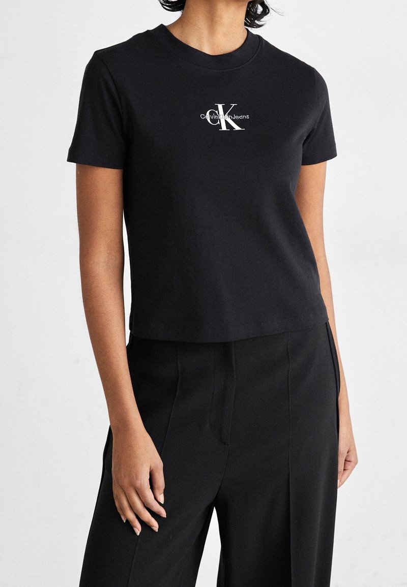 Camiseta de algodón negra corta con cuello redondo y un logo de Calvin Klein impreso en blanco en el lado izquierdo del pecho. Textura suave.
