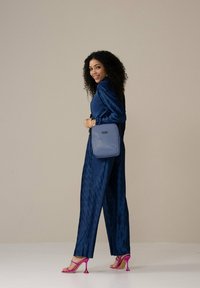 Marineblauwe plissé jumpsuit, gecombineerd met een gestructureerde grijze handtas en roze hoge hakken. De outfit heeft lange mouwen en wijde pijpen.