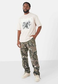 Beige grafisk t-shirt med sort print af to hoveder, kombineret med grønne camouflager cargo bukser med lommer og varierede teksturer. Casual fodtøj.