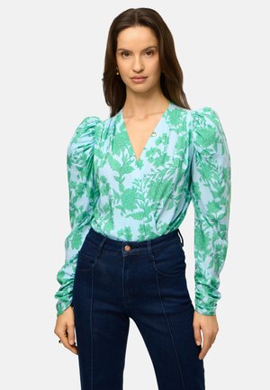 Vrouw met lang bruin haar die een lichtblauwe blouse draagt met een groene bloemendruk en poflange mouwen, gecombineerd met donkerblauwe spijkerbroek.