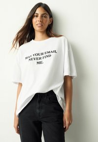 Weißes, übergroßes T-Shirt mit schwarzem Text "MÖGE DEINE E-MAIL MICH NIE FINDEN." Kombiniert mit schwarzen Jeans. Glattes Material, lässige Passform.