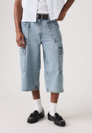 Mężczyzna w jasnoniebieskich dżinsowych krótkich spodenkach cargo, białych skarpetkach i czarnych mokasynach z frędzlami, stojący na jednolitym tle.