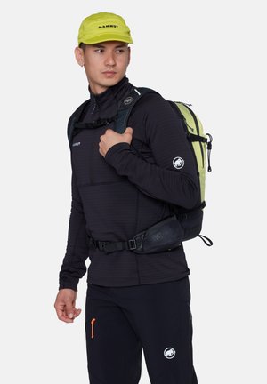 Uomo che indossa abbigliamento da esterno nero e un berretto Mammut giallo, con uno zaino da trekking verde e nero a tracolla su una spalla.