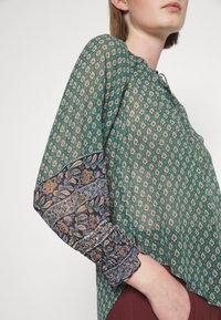 Blouse verte à motifs avec un poignet smocké et un design floral contrastant sur les manches, offrant une coupe décontractée et un col rond.