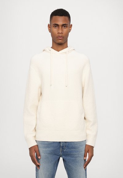 Sudadera crema con una tela texturizada, capucha con cordón y bolsillo frontal. Combinada con jeans de color azul claro que presentan un lavado desvanecido.
